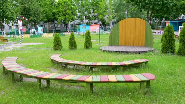 A small wooden performance stage with curved, colorful benches stands on a green grass lawn in an empty outdoor playground area surrounded by trees.
