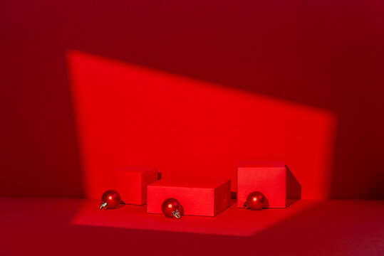 Full red monochrome holiday studio composition with a cube podium, triangular prism, scattered Christmas ornament balls, and a blank card under a diagonal light beam, suitable for product mockup.