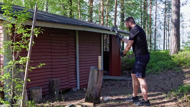 A man splits logs with an axe on a wooden chopping block outside a red wooden shed in a forest.   Horizontal at 24fps.