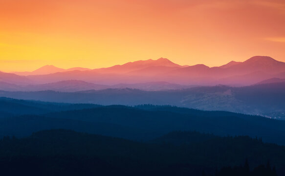 The sun sets behind the distant mountains illuminating warm orange and purple tones over the landscape. Layers of hills create a serene backdrop in the fading light.
