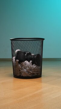 Vertical video a black digital camera and crumpled paper fill a metal mesh waste bin on a wooden floor, representing the obsolescence of technology, end of career, and environmental waste
