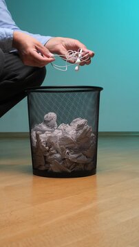 Vertical video person is responsibly disposing old tangled in ear headphones into a metal mesh waste bin filled with crumpled paper, representing electronic waste or obsolete technology