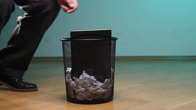 Man squatting and intentionally dropping a broken tablet and smartphone into a metal trash bin filled with crumpled paper, symbolizing digital detox, e waste, and technology obsolescence