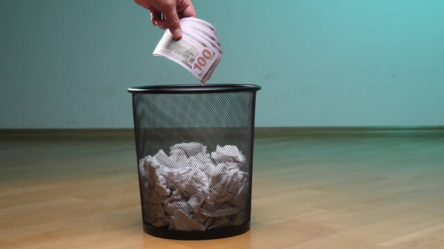 Hand placing us dollar banknotes into a trash can, symbolizing financial loss, reckless spending, waste of money, and economic downturn or poverty