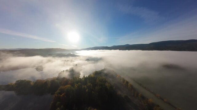 Cinematic aerial drone footage flying through dense morning fog over a peaceful mountain village with small lakes and lush forest. Low clouds cover the valley while the drone rises above the mist