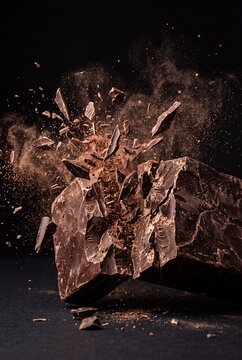 Macro shot of dark chocolate bar shattering into pieces with cocoa powder explosion on black background
