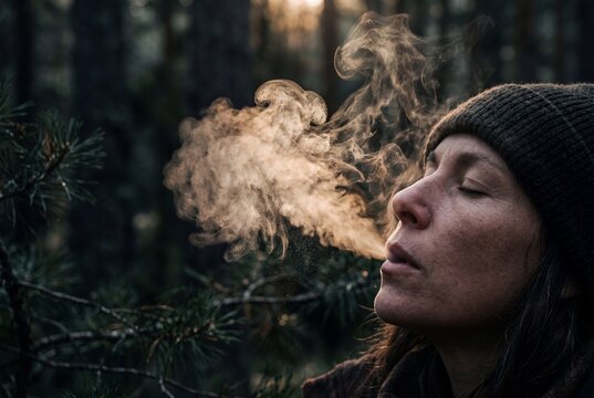 Close-up of a woman exhaling visible steam breath in a cold pine forest at sunset