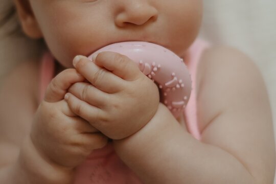 Cute baby girl in pink bodysuit lying on soft blanket and chewing silicone teether, authentic lifestyle portrait of infant teething at home.