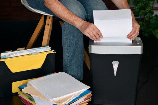 Side view mid shot showing person sitting and feeding white paper into shredder with stacks of folders and documents nearby