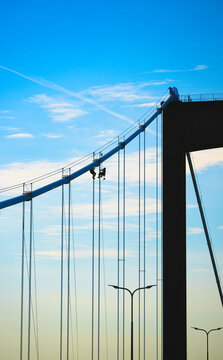 Rope access technicians inspect suspension bridge cables against blue sky.