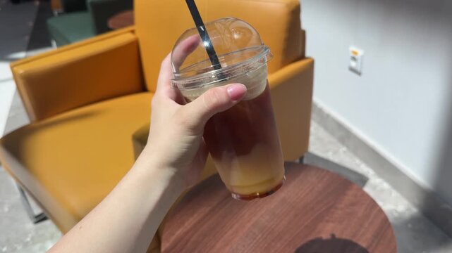 a hand holding a drink, moving it from one position to another in a casual indoor setting with yellow chairs and wooden tables, first-person perspective, neutral mood.