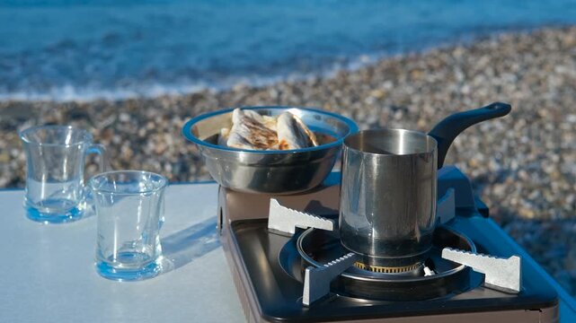 Preparing coffee and bread on portable stove at beach. Stainless steel coffee pot heating on portable gas stove on table with two empty glasses and plate of bread, on pebble beach with sea waves