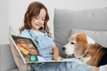 Obraz premium Cute little girl with Beagle dog reading book on sofa in living room