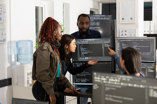 Diverse group of software developers in modern office collaborating, discussing and analyzing source code on multiple computer monitors, working together on web development project.