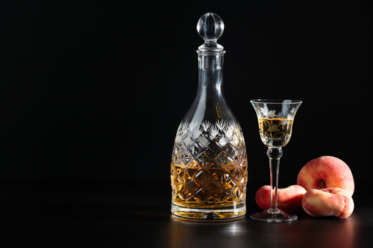 A leaded-glass carafe filled with peach liqueur and a glass are placed against a dark background. Peaches are arranged next to them. The background is dark, with space for text.