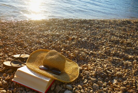 Woman woven hat , book and flip flop sandals on the beach in golden sunset, summer beach accessories