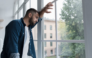 Fototapeta premium Sad young man with worried stressed face expression looking out window. Obsessive compulsive, adhd, anxiety disorders. Unhappy european handsome man stands, presses hand to glass in kitchen interior