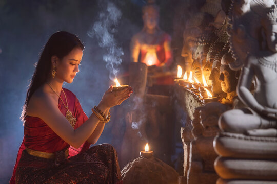 Side view of a young Thai woman in traditional clothing crouching in front of a row of Buddha statues at a temple with a candle