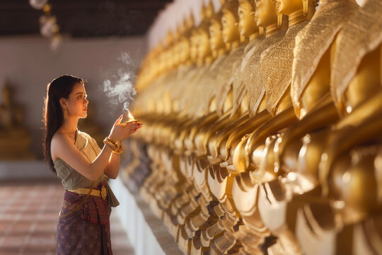 Side view of a young Thai woman in traditional clothing standing in front of a row of Buddha statues at a temple holding a candle