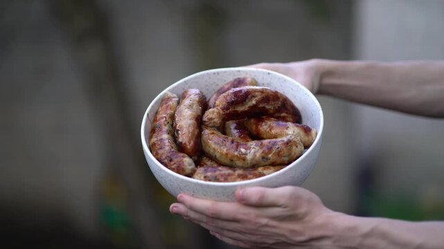 Golden brown grilled sausages with char marks in speckled bowl held by hands. Appetizing bbq food perfect for summer picnics and outdoor cooking videos.