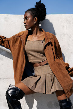 Profile view of a stylish young Black woman in a checkered mini skirt and brown jacket posing against a concrete wall