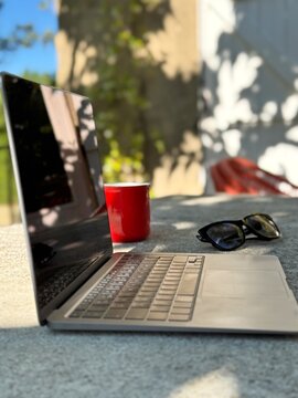 Close-up side view of an open laptop computer with a blank screen on a garden table with a pair of sunglasses and a mug on a summer day