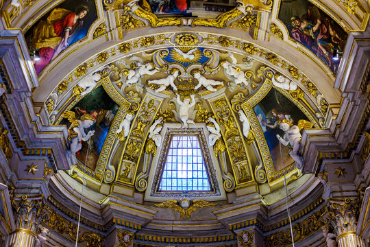 Ornate ceiling inside the Duomo di Urbino, Italy, featuring a central dove and cherub reliefs surround a clear window, complementing vibrant religious paintings and rich, gilded rococo decorations