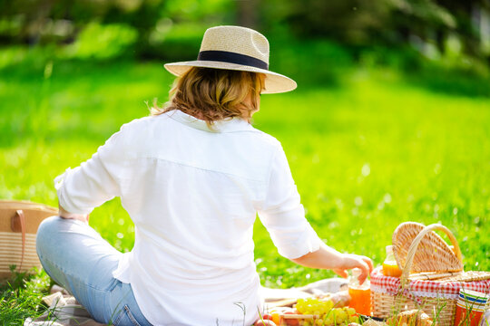 Adult woman in white shirt and straw hat seen from behind reaching for juice bottle. Outdoor hydration refreshment recreation concept.