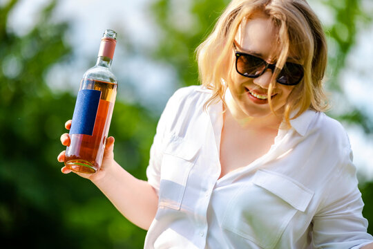 Smiling woman in sunglasses holding a rose wine bottle, outdoors on a sunny day. Summer picnic relaxation and cheerful lifestyle concept.