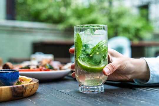 Close-up side view of a woman sitting at a table at an outdoor cafe terrace holding a refreshing mojito cocktail