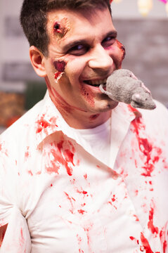 Man dressed as a bloody zombie holds a rat in his mouth during a Halloween celebration, creating a disturbing horror costume moment.