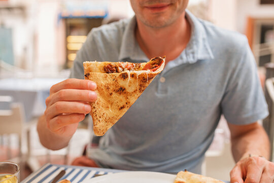 Close-up of a smiling man sitting at an outdoor restaurant eating a slice of prosciutto ham and aubergine pizza