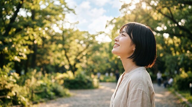 ショートヘアの若いアジア人女性が、公園の緑豊かな木々から差し込む温かい日差しを見上げながら明るく微笑む