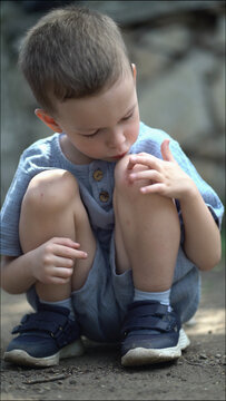 A cute child examines his knee wound outdoors by blowing on it and touching it gently. Sad expression in this authentic moment with natural daylight.