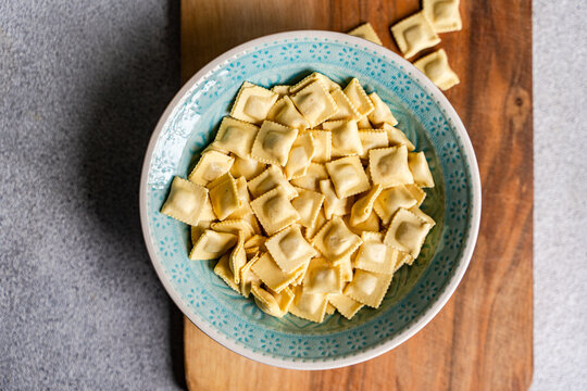 Close-up overhead view of a bowl of uncooked ravioletti pasta on a wooden chopping board