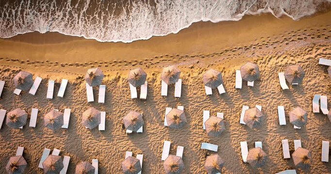 Aerial top view over beach umbrellas and sunbeds on golden sand coastline and sea waves washing the shore at sunset summer vacation