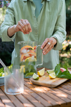 Woman standing in a garden using a long spoon to stir assorted citrus fruits and mint in a glass pitcher of water