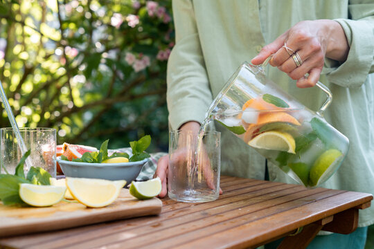 Close-up of a Woman standing by a garden table pouring a glass of fresh water with assorted citrus fruit and mint leaves