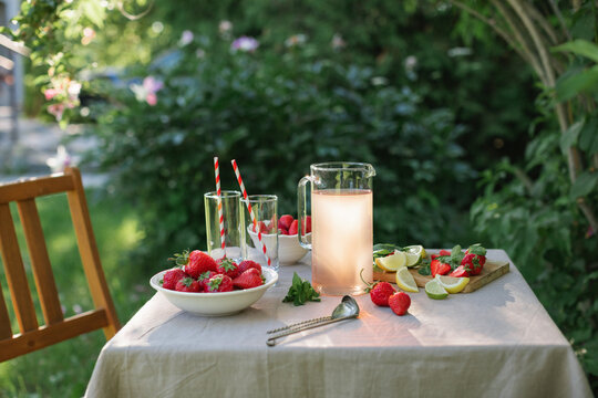 Close-up of a pitcher of strawberry lemonade, two glasses, fresh strawberries, limes and mint leaves on a garden table on a summer day
