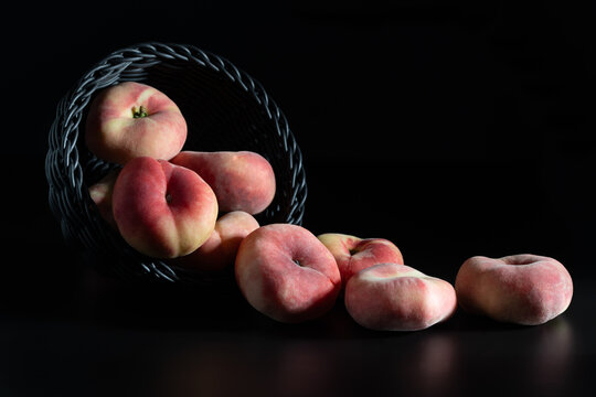 Close-up of fresh flat peaches falling out of a basket. The basket and the background are dark. the red peaches provide a contrast.