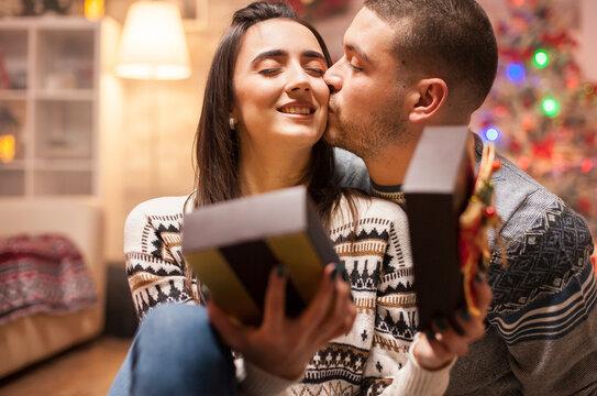Happy boyfriend kisses his girlfriend cheek after giving her a Christmas gift, creating a tender romantic holiday moment.