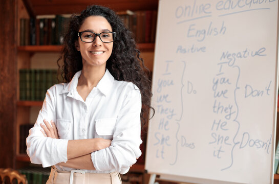 Learning And Knowledge Concept. Portrait of proud smiling woman in eyeglasses posing near whiteboard with English language grammar rules, confident positive teacher lady standing with folded arms