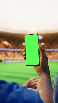 Vertical composition: Close-up of mockup smartphone used by man in soccer stadium stands to compare match odds, with football supporter checking live score application predictions on chroma key phone.