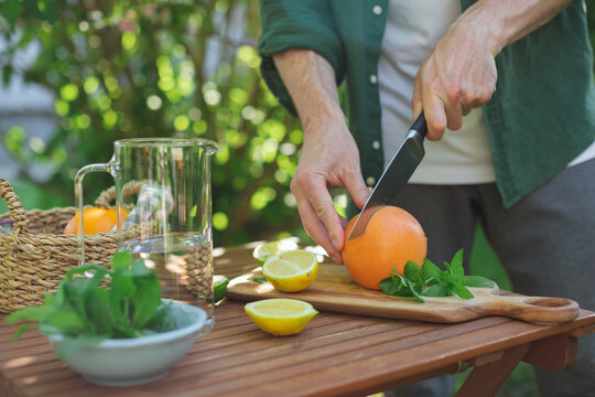 Close-up of a man standing by a garden table cutting a fresh grapefruit to make a citrus lemonade drink on a summer day