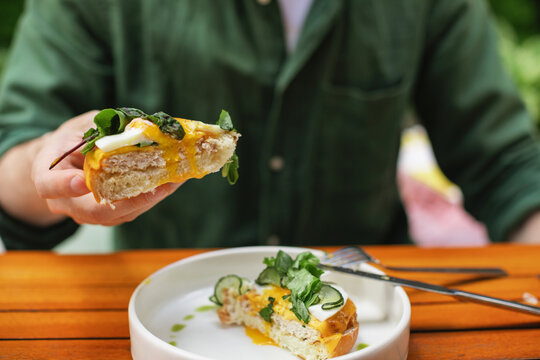 Close-up of a man sitting at a table eating a halved open-faced toasted egg and cheese bun