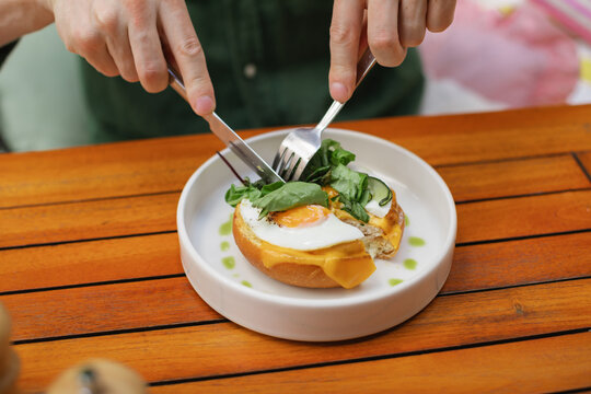 Close-up of a man sitting at a table eating an open-faced toasted egg and cheese bun with a knife and fork