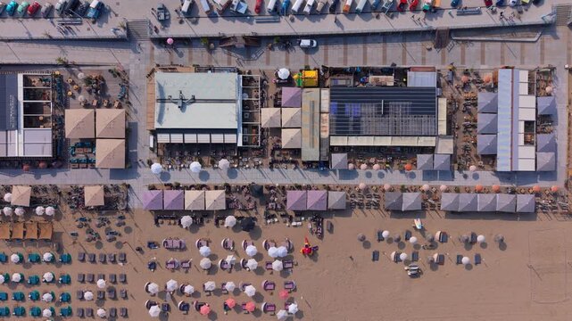Aerial top down view of multiple modern beach clubs with outdoor restaurant terraces large sun umbrellas solar panels and neat rows of lounge chairs on a wide sandy resort beach