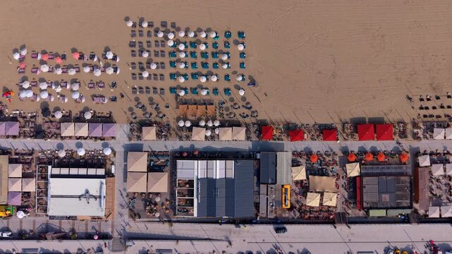 Aerial top down view of a vibrant coastal strip with seasonal beach clubs outdoor dining terraces colorful sun umbrellas and geometric rows of sunbeds on a wide sandy beach