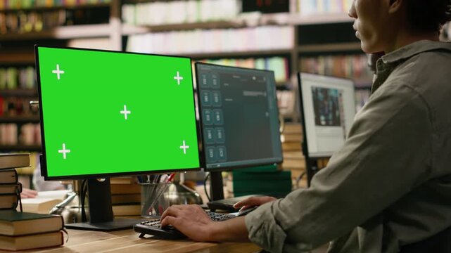 Green screen on desktop monitor near asian student typing at table, using mockup screen replacement on computer display, library workstation with books, for editable screen mockup and VFX use.
