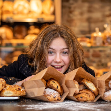 Excited young girl smiles brightly, peeking over a counter adorned with multiple paper bags brimming with fresh, sweet bakery treats.